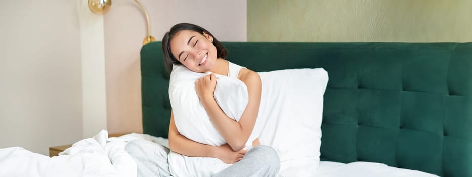 Romantic smiling asian girl lying in her bed, hugging pillow and gazing dreamy, lazy mornings in bedroom.