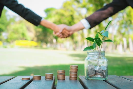 Sustainable money growth investment, glass jar filled with money saving and coin stack with businesspeople handshake in background as eco-friendly financial investment nurtured with nature. Gyre