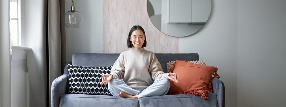 Relaxation and patience. Smiling young asian woman in cozy room, sitting on sofa and meditating, doing yoga mindfulness training.