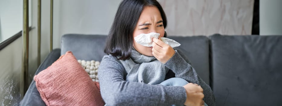 Close up of korean girl sneezing in napking, sitting at home in scarf, feeling unwell, concept of winter, heating cost and people.