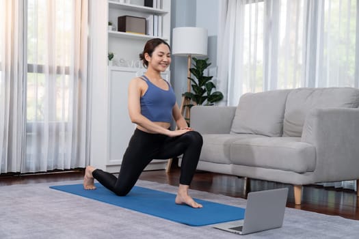 Asian woman in sportswear doing yoga exercise on fitness mat as her home workout training routine. Healthy body care lifestyle woman watching online yoga video on laptop. Vigorous