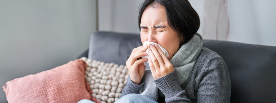 Close up of young korean woman staying at home with cold, sneezing in napking, has runny nose, concept of illness, health and influenza.