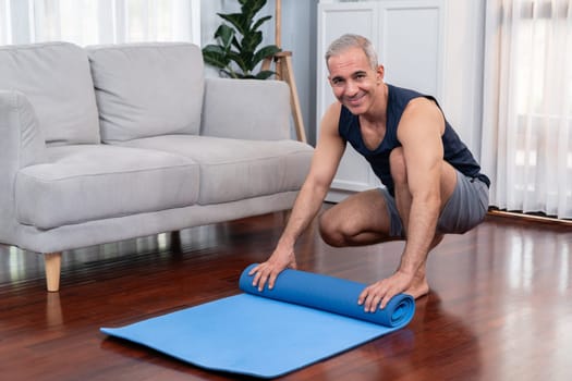 Active and sporty senior man preparing, rolling fitness exercising mat on living room floor at home. Home exercise as concept of healthy fit body lifestyle after retirement. Clout