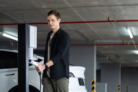 Young man travel with EV electric car to shopping center parking lot charging in downtown city showing urban sustainability lifestyle by green clean rechargeable energy of electric vehicle innards