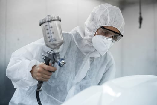 Automotive service worker in full protective gear expertly apply color paint in to car's bodywork with spray gun or respirator painting in chamber workshop. Car paint service for scratch refinish.Oxus