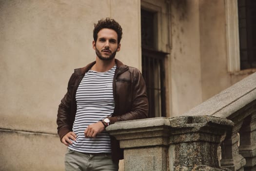 A man in a striped shirt and leather jacket leaning on a railing and looking at camera