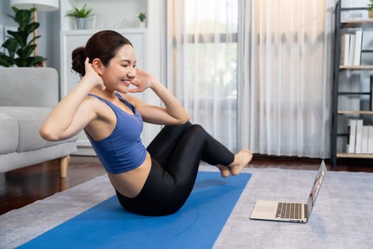 Asian woman in sportswear doing crunch on exercising mat as home workout training routine. Attractive girl engage in her pursuit of healthy lifestyle with online exercise training video. Vigorous