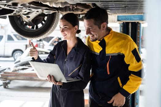 Two vehicle mechanic working together underneath lifted car, conduct car inspection with laptop. Automotive service technician in uniform carefully make diagnostic troubleshooting. Oxus