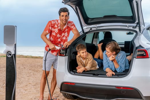 Family vacation trip traveling by the beach with electric car, lovely family sit on the trunk, charging EV car battery with green and sustainable energy. Family travel and eco-friendly car. Perpetual