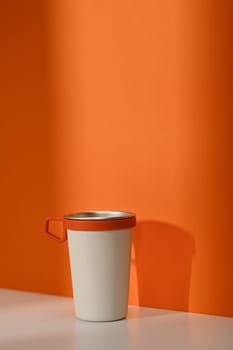 A white travel cup with handle over orange wall. Tumbler beverage container and Eco friendly lifestyle.