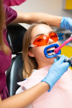 Woman wearing protective goggles from ultraviolet light sits with tube in mouth to absorb liquid. Dentist places tube in patient mouth and works on teeth