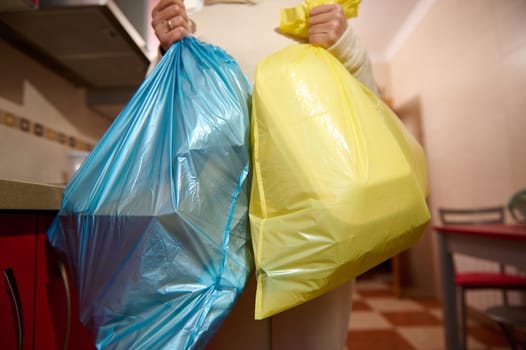 A person carrying blue and yellow garbage bags while standing in a brightly lit kitchen. The bags appear full, symbolizing domestic activities like cleaning or waste disposal.