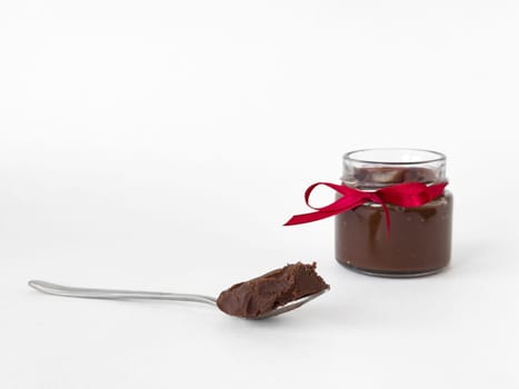 Elegant glass jar of hazelnut chocolate spread with red ribbon and spoonful on white background.