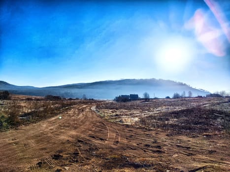 A dirt road winds through an open landscape at dawn, with a hillside and bright blue sky in the background.