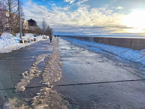 A peaceful walk takes place along the embankment in Arkhangelsk as spring brings melting snow and a vibrant sky filled with clouds.