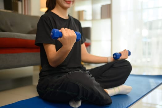 Young woman doing exercise with dumbbells on mat in her living room..