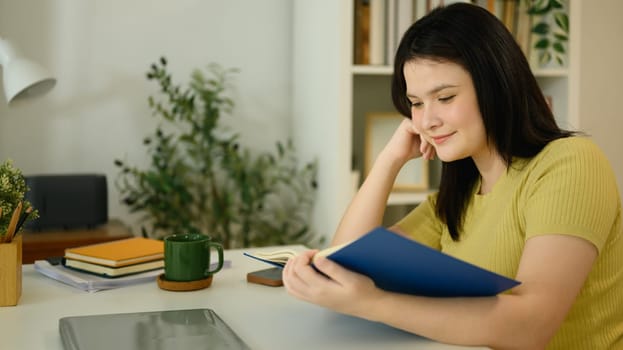 Calm young woman reading book at desk in her living room.