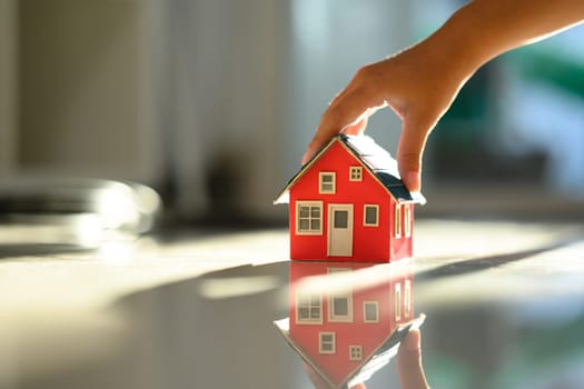Hand placing a small house model on reflective surface with soft, natural light. Real estate and housing aspirations concept.