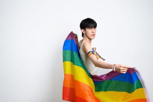 Portrait of young man holding rainbow flag against white background, celebrating LGBTQ pride.