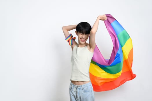 Portrait of young man holding rainbow flag against white background, celebrating LGBTQ pride.