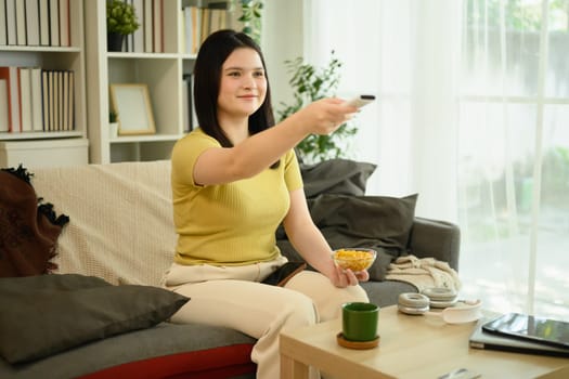 Beautiful young woman holding a bowl of cereal and remote control watching television in her living room.