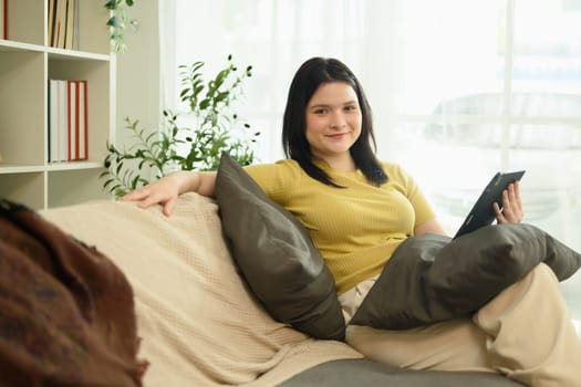 Beautiful young woman in casual clothing using digital tablet on couch in bright living room.