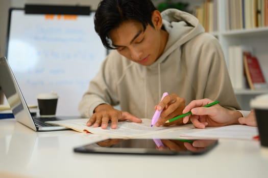 Focused male student highlighting important text with a marker while studying with his tutor.