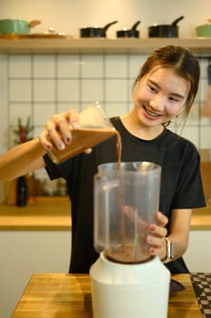 Smiling young woman preparing a healthy drink pouring chocolate liquid into a blender.