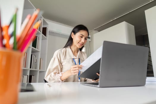 A young businesswoman reviews financial documents in a modern office, focusing on her work with a laptop and paperwork.