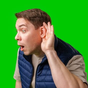 Surprised young man holding his hand to his ear, listening intently against a vibrant green background in studio