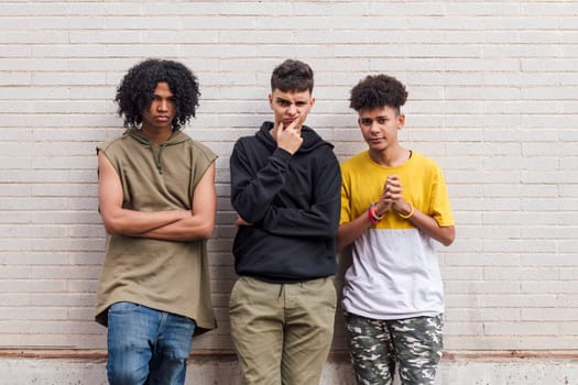 three multiethnic teenager friends with folded hands leaning against white brick wall, concept of friendship in youth and urban lifestyle
