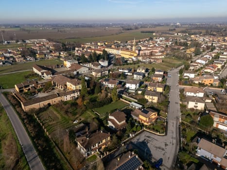 Aerial view capturing the small town of Stagno Lombardo in Cremona, Italy, featuring residential houses with solar panels, a church, and cultivated fields under a clear blue sky