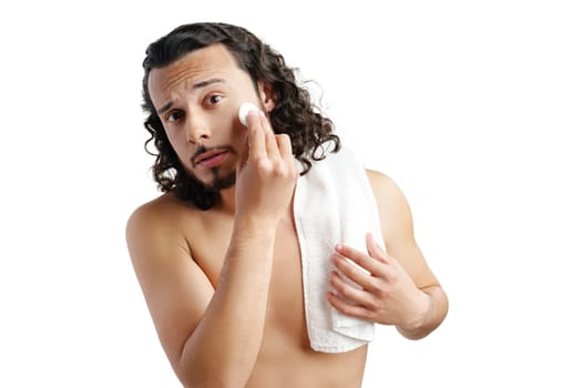 A young man with curly hair applies a skincare product to his face in a well-lit space close up