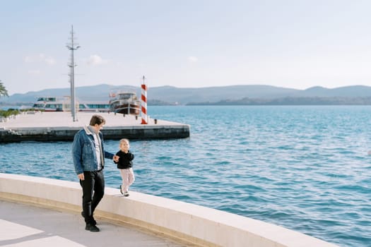 Dad leads a little girl by the hand along a low fence along the boardwalk. High quality photo