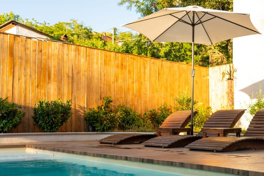 Backyard poolside with wooden sun loungers and umbrella. Cozy outdoor area bathed in golden sunlight