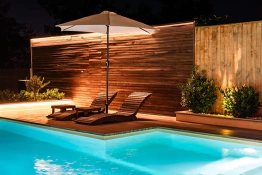 Elegant swimming pool with lounge chairs and umbrella at night. Luxury outdoor space illuminated with warm lights