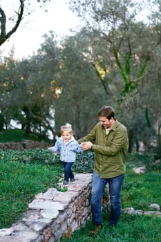 Smiling dad leads a little girl along a stone fence in the park walking next to the grass. High quality photo