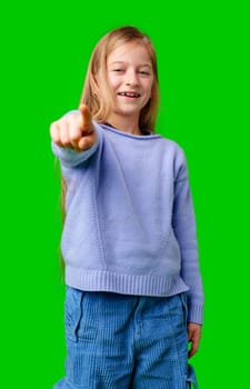 A young girl in a lavender sweater smiling and pointing at the camera against a bright green background in studio