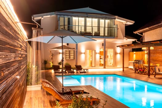 Luxury villa featuring an illuminated swimming pool, wooden deck, and cozy ambient lighting at night. Concept of a stylish and modern outdoor living space for comfort and relaxation