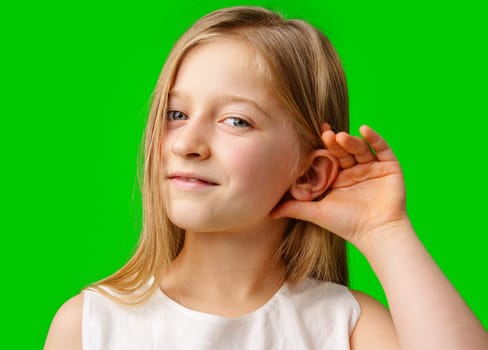 A young girl with long blonde hair cupping her hand to her ear against a bright green background in studio