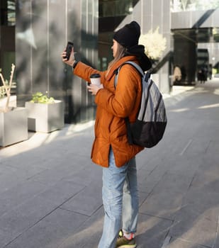 A young individual happily captures a cheerful selfie while holding a warm cup of coffee in a lively urban environment