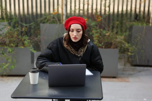 A young woman in a red beret works on her laptop at an outdoor caf, enjoying her coffee