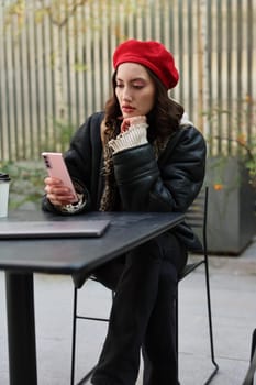 A stylish young woman wearing a vibrant red beret is absorbed in her phone, showing off trendy urban fashion vibes