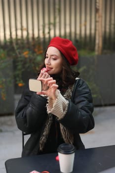 A stylish young woman wearing a vibrant red beret enjoys sipping her coffee while taking a fun selfie outdoors
