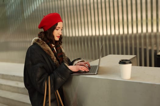 A stylish and fashionable young woman wearing a striking red beret is using her laptop outdoors while sipping coffee