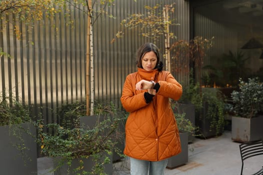 A woman in a bright orange coat checks her watch surrounded by lush greenery in a modern outdoor space