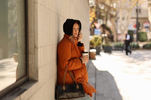 A woman in a bright orange coat holds a warm coffee beside a wall as the busy city vibrantly moves around her