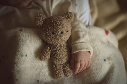 Fluffy toy teddy bear and baby little hand lying in bed in warm, cozy sunlight. Childhood and parenting concept. High quality photo