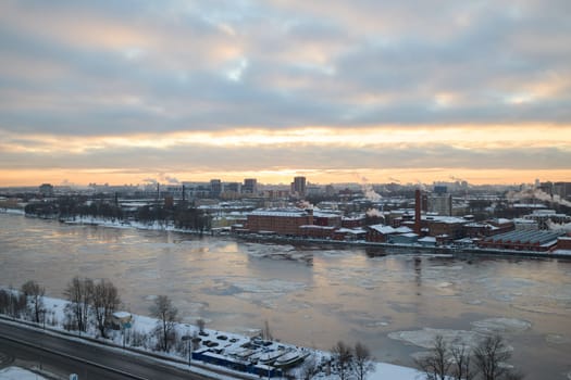 A beautifully serene winter view depicts a river covered in snow alongside urban buildings during a tranquil sunset