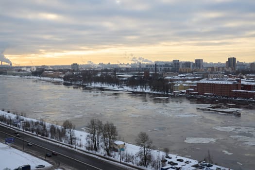 A serene winter view of a tranquil river, surrounded by snow, buildings, and a cloudy sky at sunset time